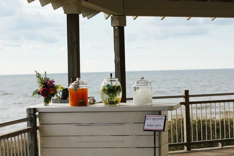 A beverage station with three large glass jars filled with colorful drinks and ice, set on a white wooden counter with a small bouquet of flowers, overlooking the ocean on a cloudy day.