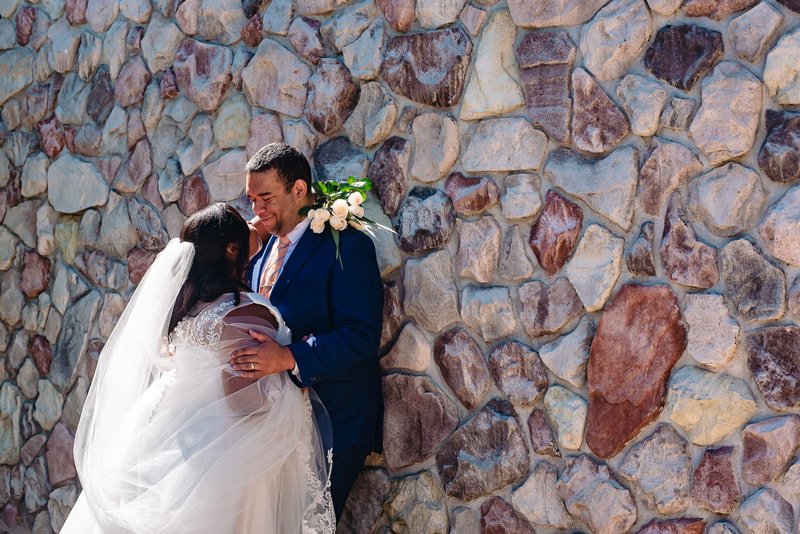 Bride and groom sharing a moment in front of a stone wall, bride in a white dress and veil, groom in a blue suit with a boutonniere.
