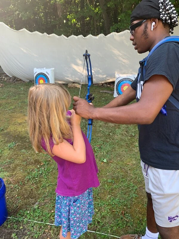 A young girl with long blonde hair is aiming a bow and arrow while an adult man assists her at an outdoor archery range. There are two targets with colorful concentric circles in the background, and a large white curtain or sheet behind them, set up 