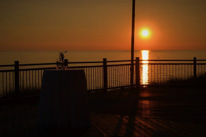 Sunset over the ocean viewed from a wooden deck with a table and a railing.