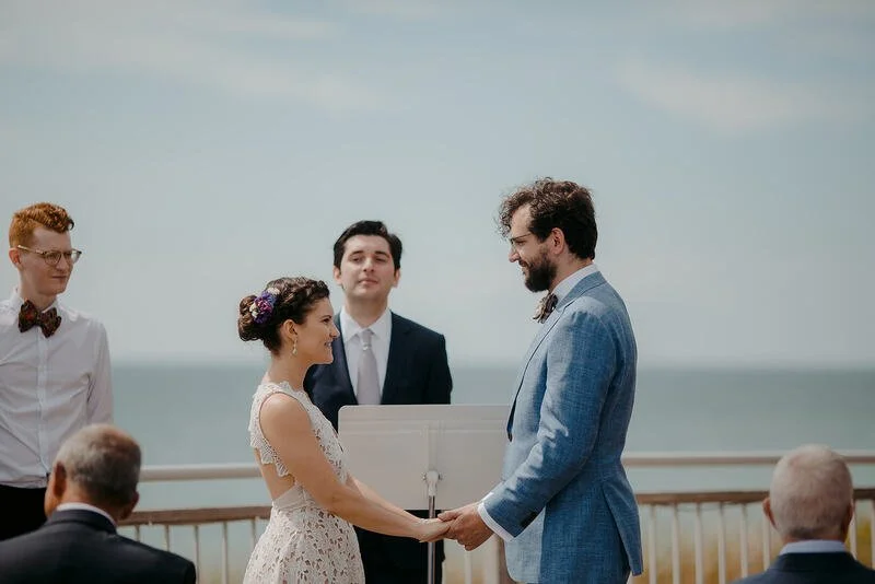 A couple getting married on an outdoor deck overlooking the ocean, with officiant and guests present.