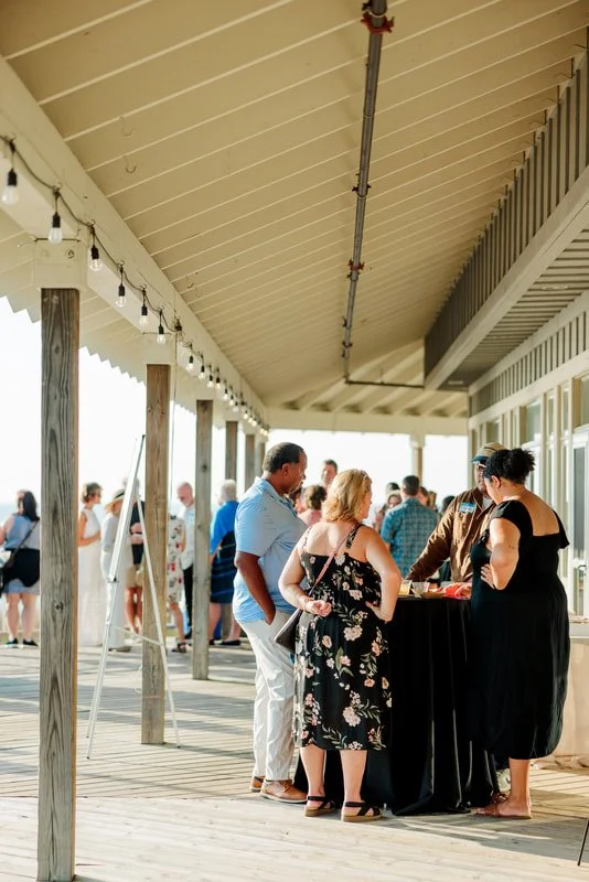 People gathered around a table at an outdoor event or gathering, on a covered wooden deck. Some are talking and some are walking around in the background.