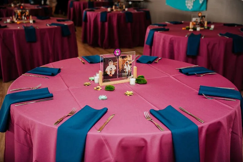 Round table with pink tablecloth, set for dining with blue napkins and silverware, central decorative photo frame, surrounded by other similarly decorated tables at a party or event.
