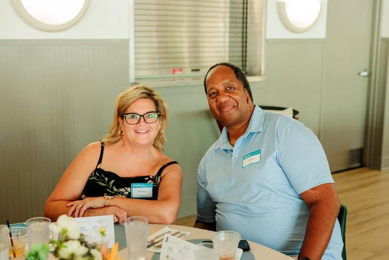 A woman and a man sitting at a table during a social event or conference in a well-lit room, smiling at the camera.