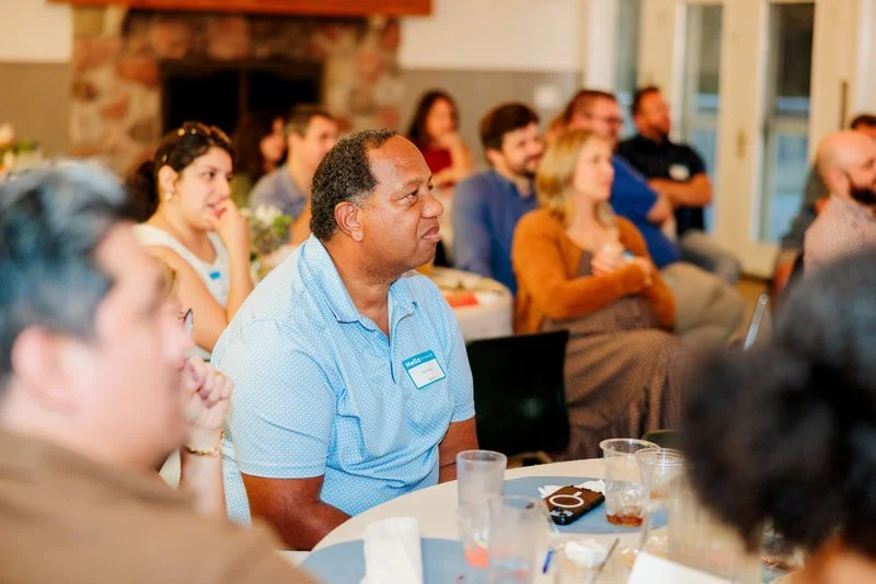Group of people attending a meeting or seminar, sitting at tables in a room with a fireplace in the background.