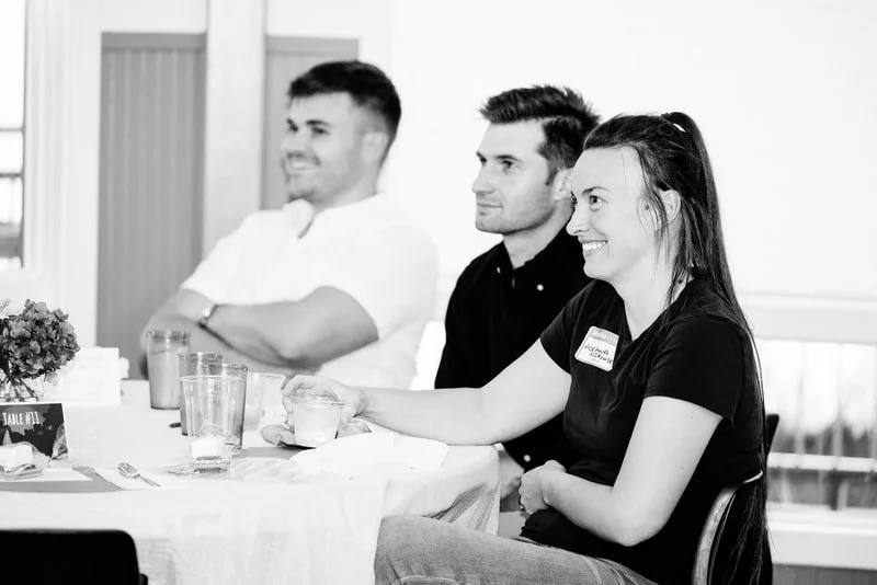 Three people sitting at a table during a meeting or event, smiling and engaging with others.