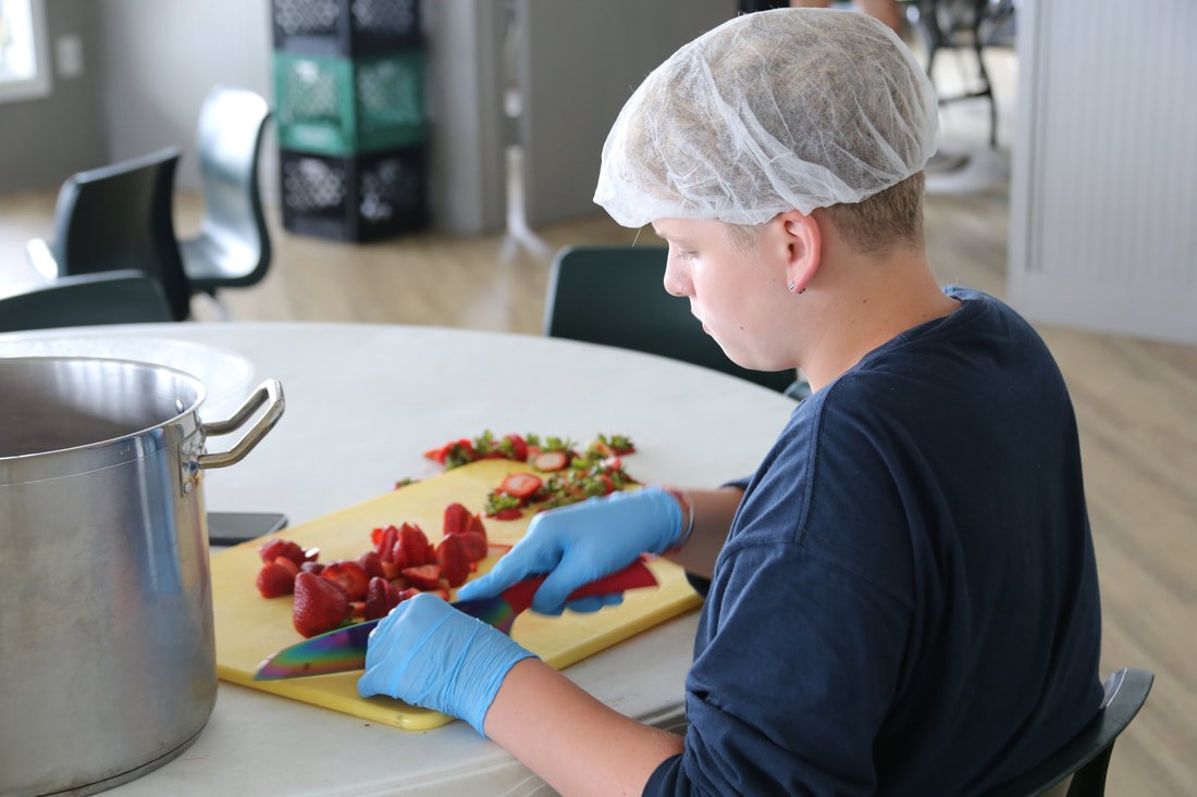 A young person wearing a hairnet and blue gloves chopping strawberries on a yellow cutting board in a kitchen or cafeteria.