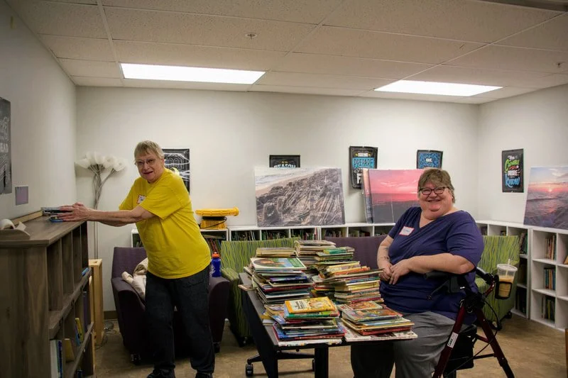 Two women organizing books in a brightly lit room with artwork on the walls and shelves filled with books.
