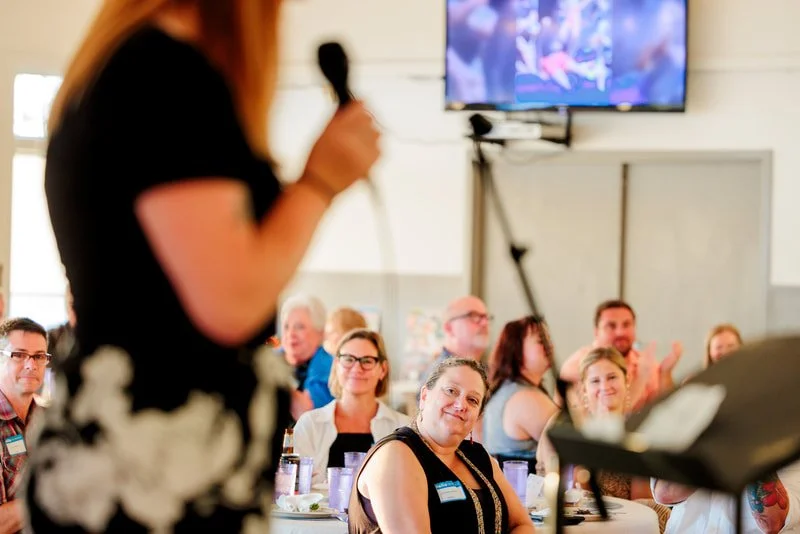 A woman with long blonde hair speaking into a microphone at a conference or seminar, facing an audience of diverse people applauding. There is a television screen mounted on the wall showing a colorful scene.