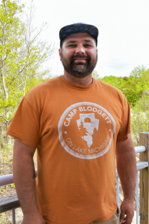 A man with a beard and baseball cap smiling outdoors wearing an orange Camp Blodgett T-shirt with a white graphic of Michigan.