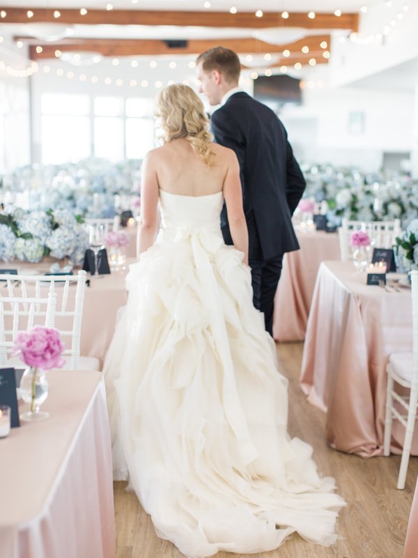 Bride and groom walking at their wedding reception, with tables decorated with pink and white flowers and string lights overhead.