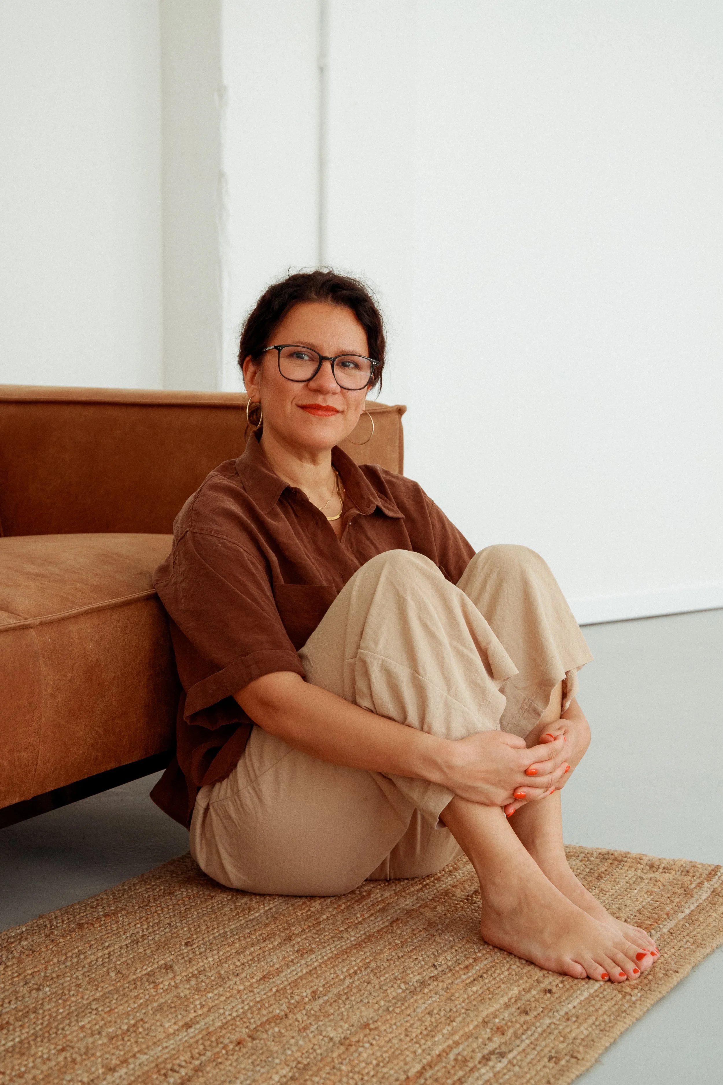Eine Frau sitzt auf einem braunen Sofakissen vor einer weißen Wand, trägt eine braune Bluse, beige Hosen, Gläser und hat dunkle, kurze Haare, entspannt mit verschränkten Armen