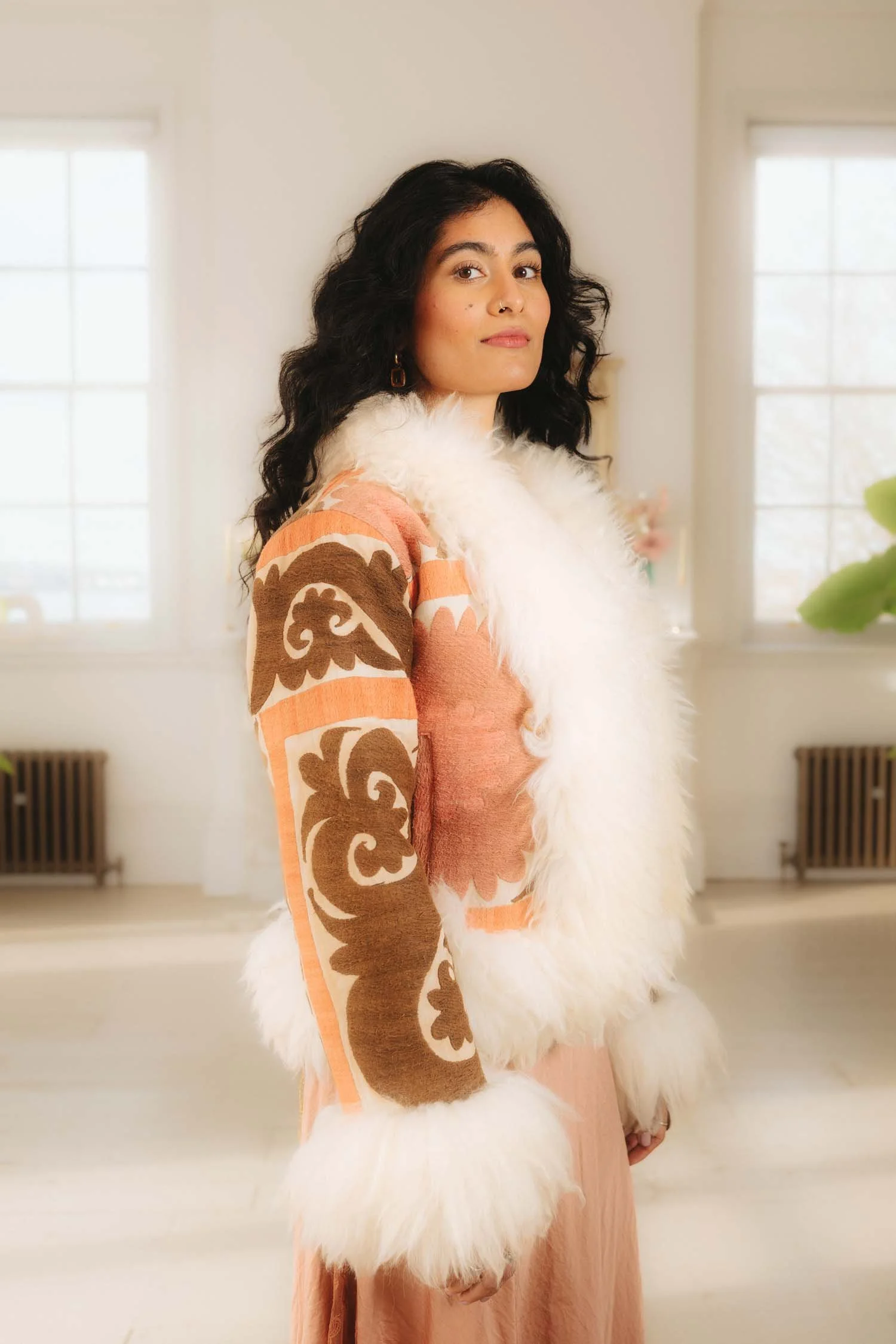 A woman with dark, curly hair wearing a pink and brown patterned coat with white fur trim, standing in a brightly lit room with large windows.