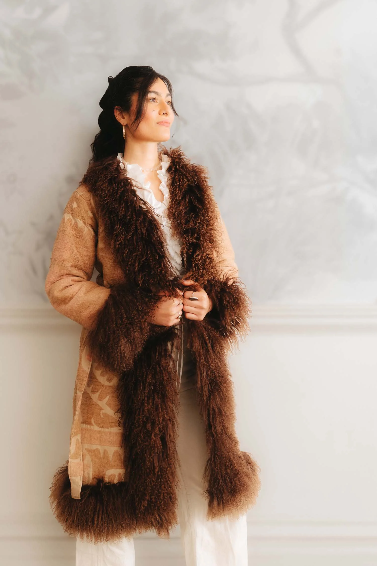 A woman standing against a light gray wall, wearing a long coat with brown fur trim and a white ruffled blouse.