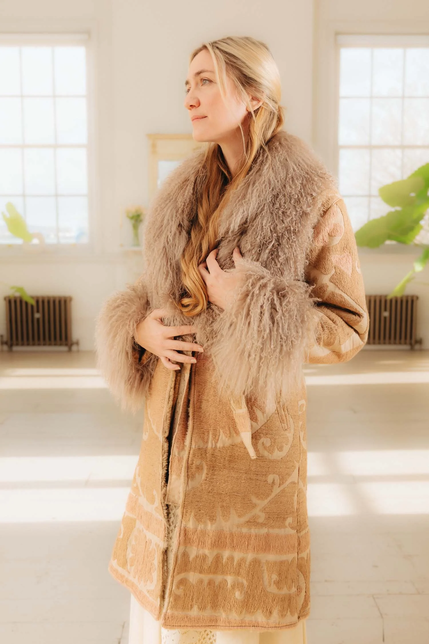 A woman with long blonde hair wearing a beige suzani coat, with taupe mongolian fur trimming the edges, standing indoors in front of windows with sunlight, holding her coat closed with a gentle expression.