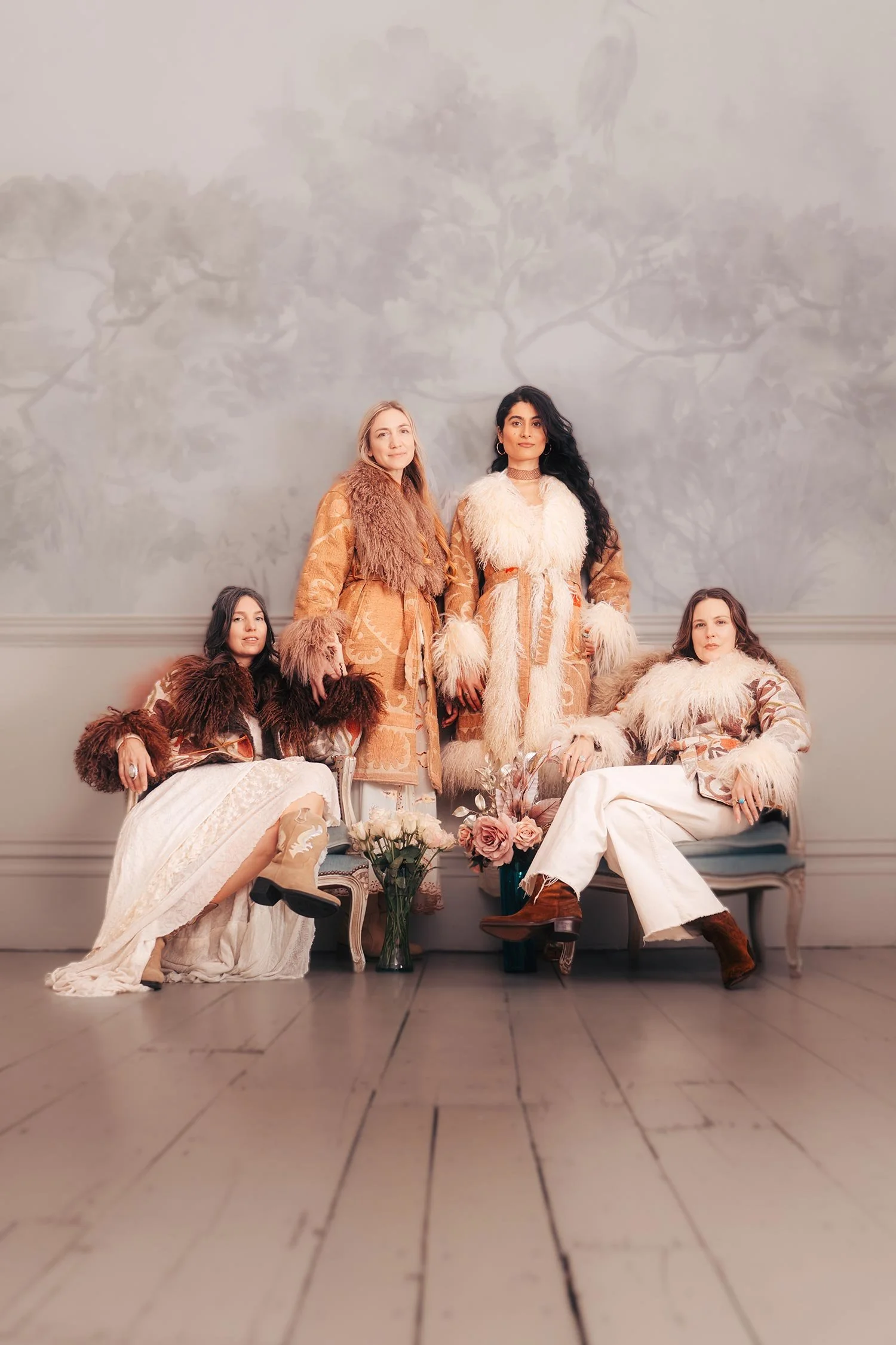 Four women dressed in fashionable, vintage-inspired outfits wearing vintage suzani fabric coats with fur accents, sitting and standing on a sofa in a room with wooden floors and a painted wall mural of trees, with roses and flowers by chairs