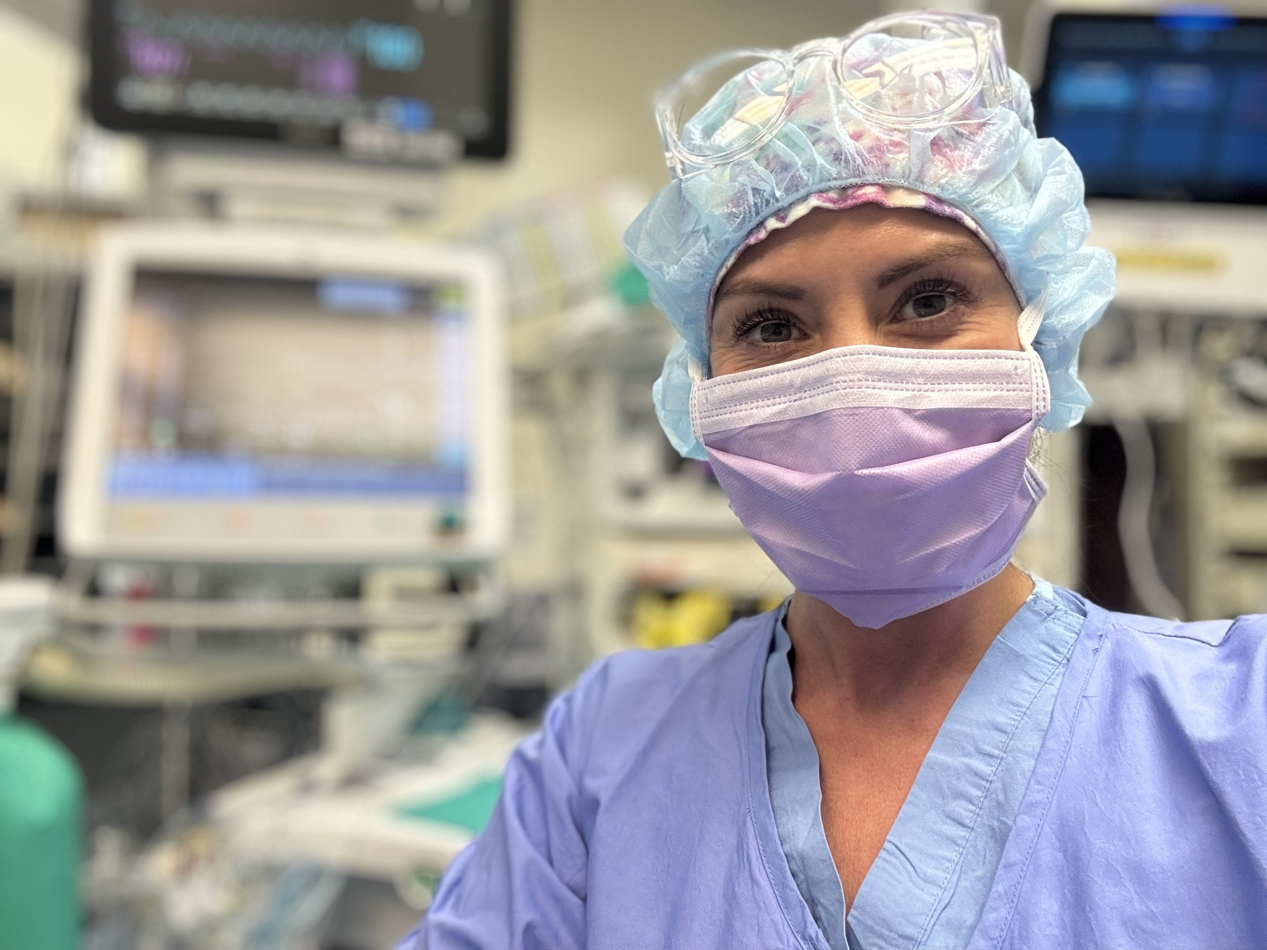 A female healthcare worker in scrubs, wearing a purple face mask, surgical cap, and glasses on top of her cap, in a medical setting with monitors and medical equipment in the background.