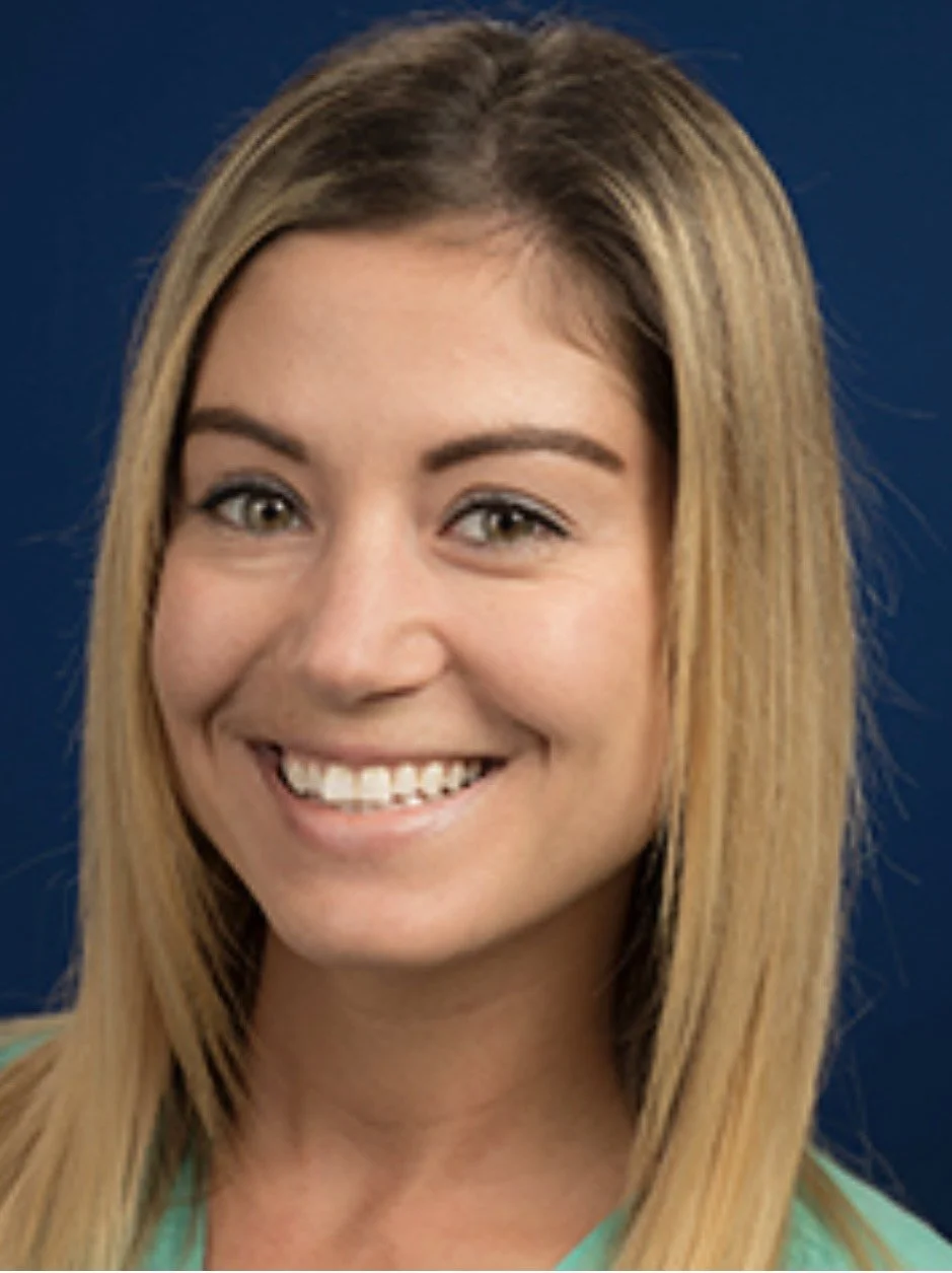 Close-up of a smiling woman with blonde hair and light skin, wearing a green top, against a dark blue background.