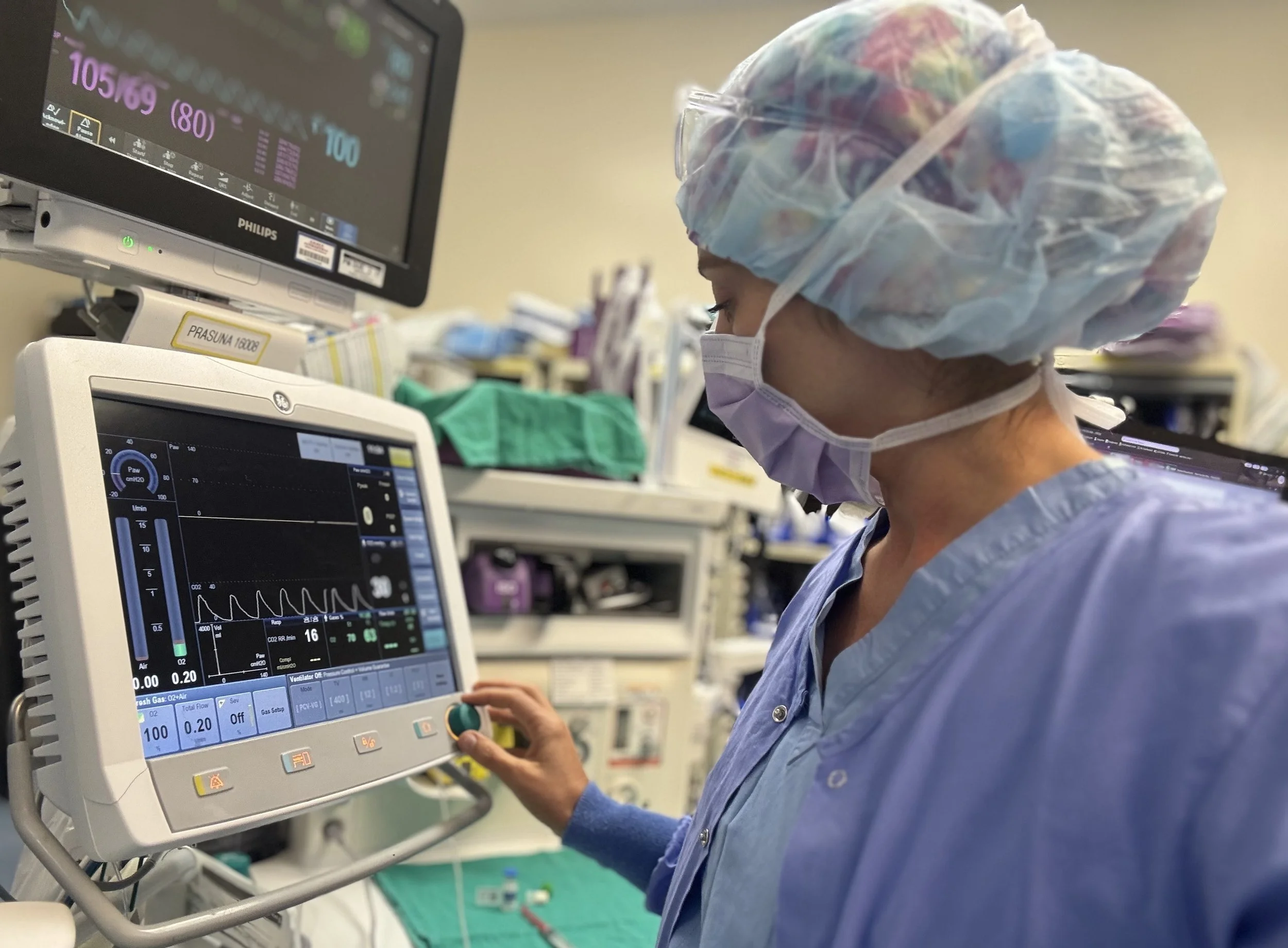A healthcare worker in scrubs, a surgical cap, and a face mask operates a medical ventilator in a hospital setting.