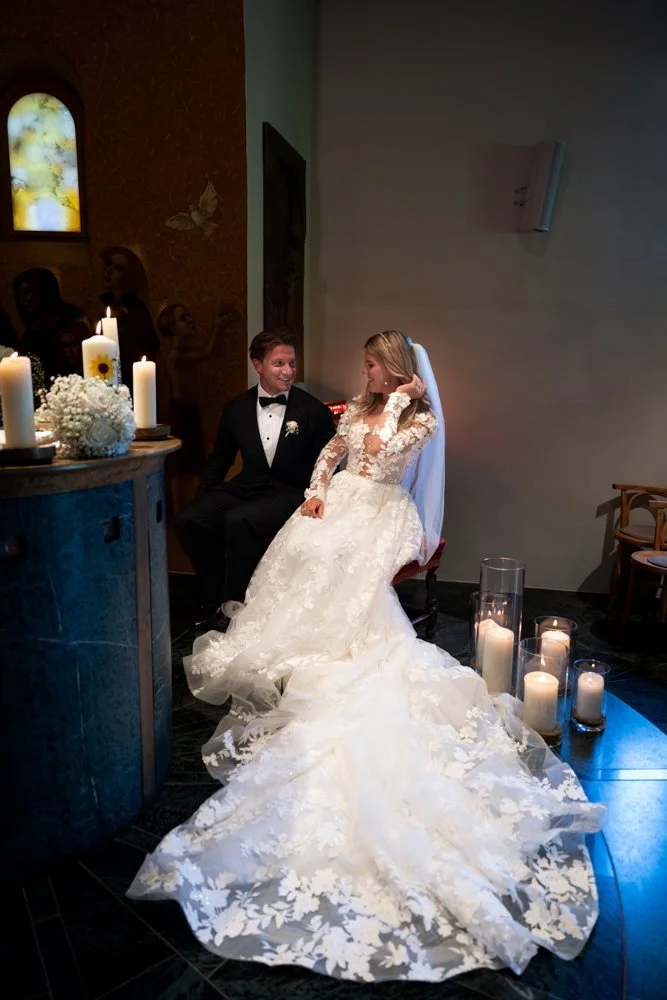 Ein Brautpaar sitzt zusammen bei einer Hochzeit, die Braut trägt ein langes weißes Hochzeitskleid mit Spitzenmuster, der Bräutigam trägt einen schwarzen Anzug mit Fliege.