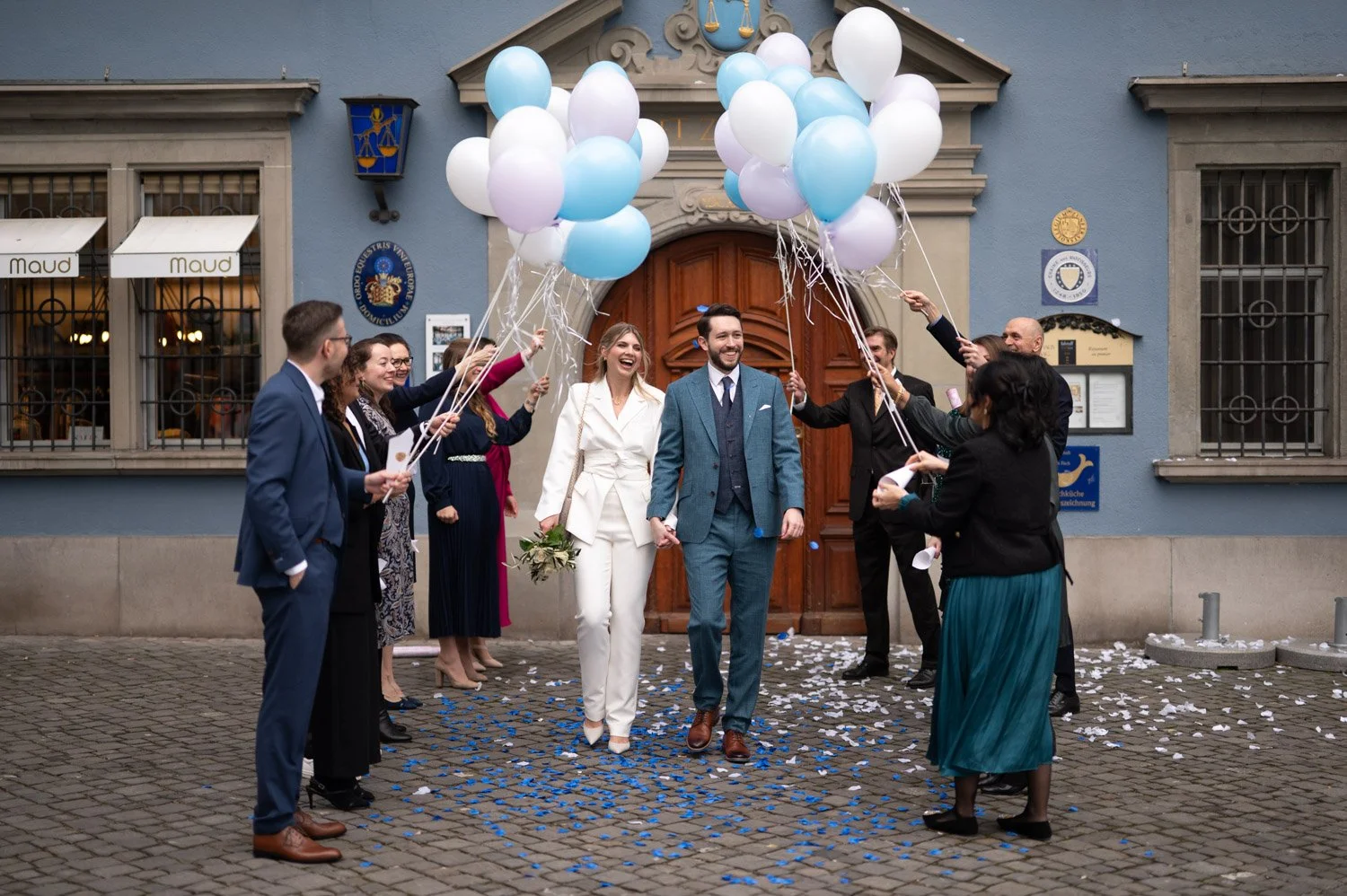 Ein frisch vermähltes Paar wird an der Eingangstür eines Gebäudes von Freunden und Familie mit Luftballons und Konfetti gefeiert.