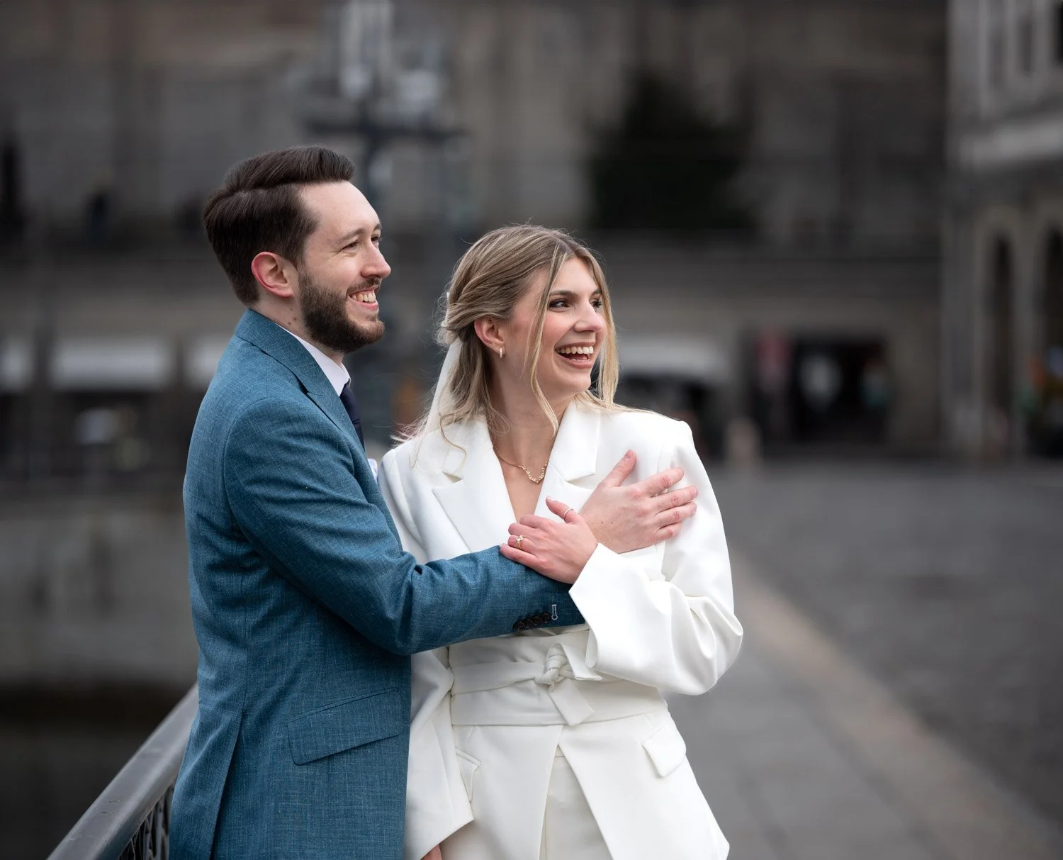 Ein lachendes Paar in formeller Kleidung steht im Freien auf einer Brücke und umarmt sich.