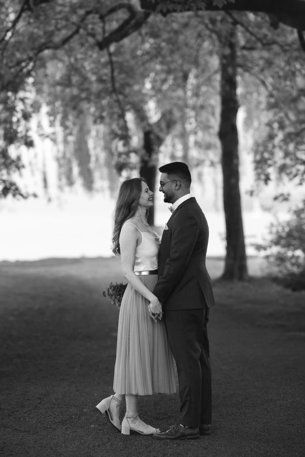 Ein Paar hält Händchen und blickt sich verliebt an, in einem Park mit Bäumen, schwarz-weiß Fotografie.