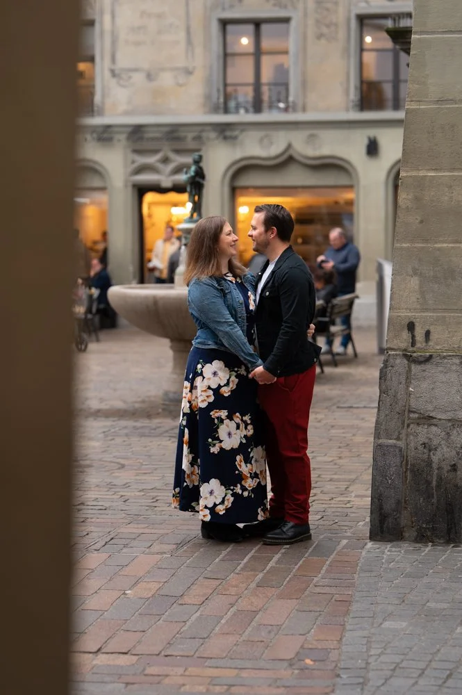 Ein verliebtes Paar hält Händchen in einer belebten, historischen italienischen Stadt mit terrakottafarbenen Gebäuden und einem Brunnen im Hintergrund.
