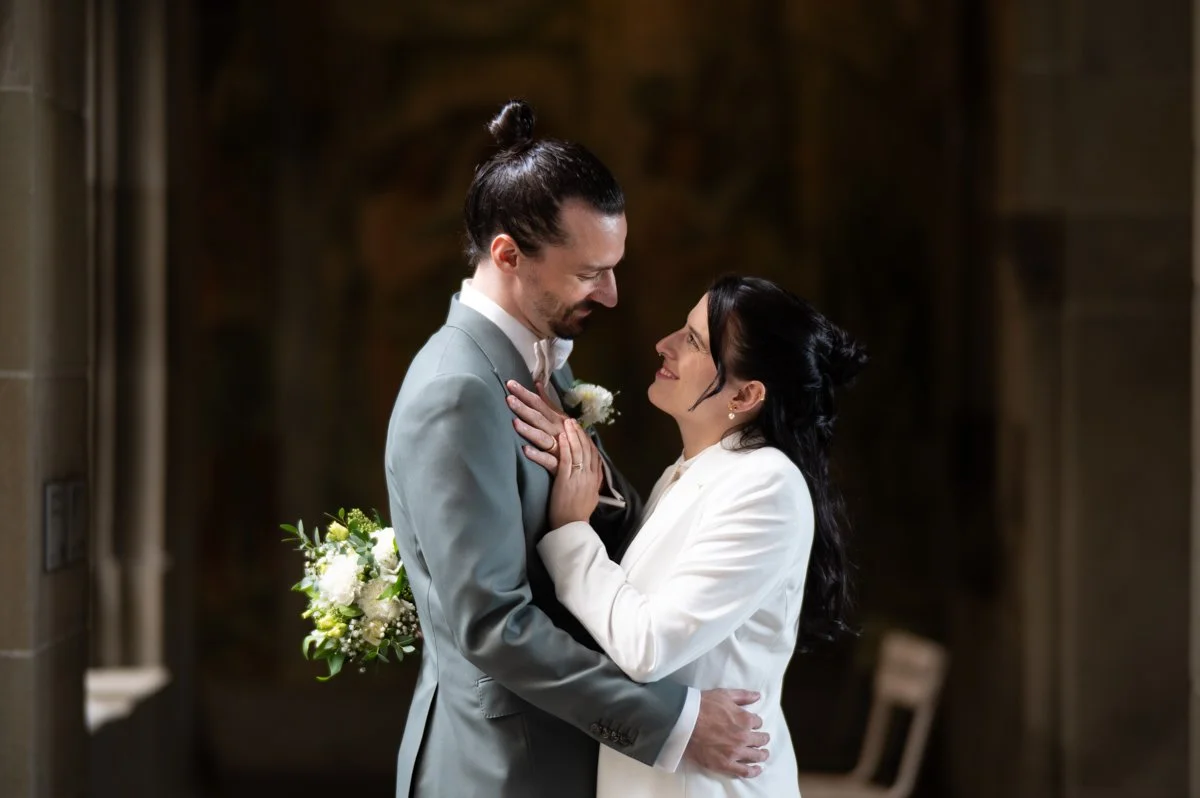 Braut in weißem Kleid und Bräutigam in grauem Anzug bei Hochzeitszeremonie, die sich liebevoll umarmen.