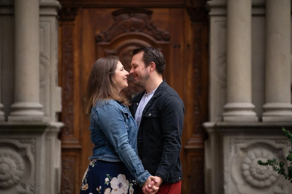 Ein Paar hält Händchen und schaut sich verliebt an vor einer kunstvollen Holztür mit steinernen Säulen im Hintergrund.