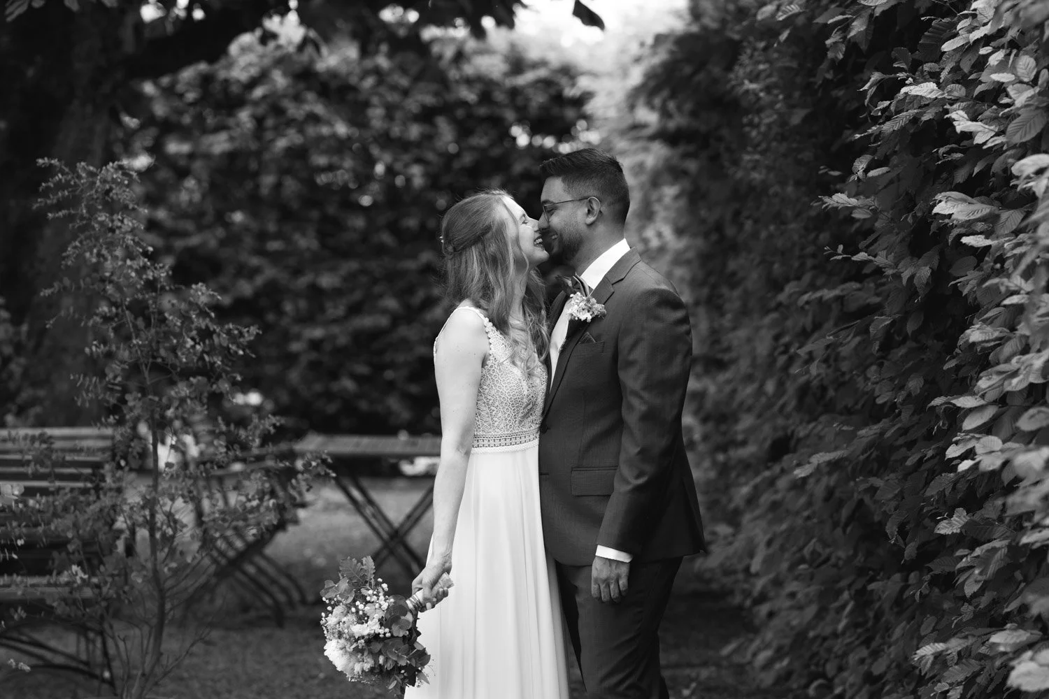 Ein schwarzes und weißes Hochzeitsfoto von einem Paar, das sich liebevoll umarmt und küsst, im Freien vor einer dichten Hecke, mit einem Blumenstrauß in der Hand der Braut.