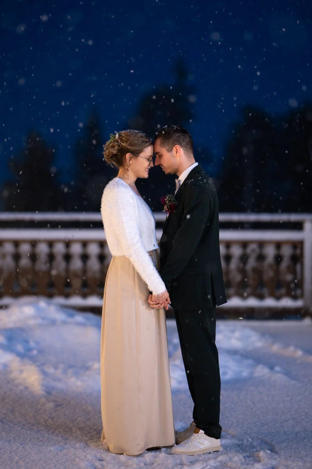 Ein Brautpaar steht sich im Schnee gegenüber mit den Stirn aneinander, in einem Moment der Intimität während einer Winterhochzeit am Abend.