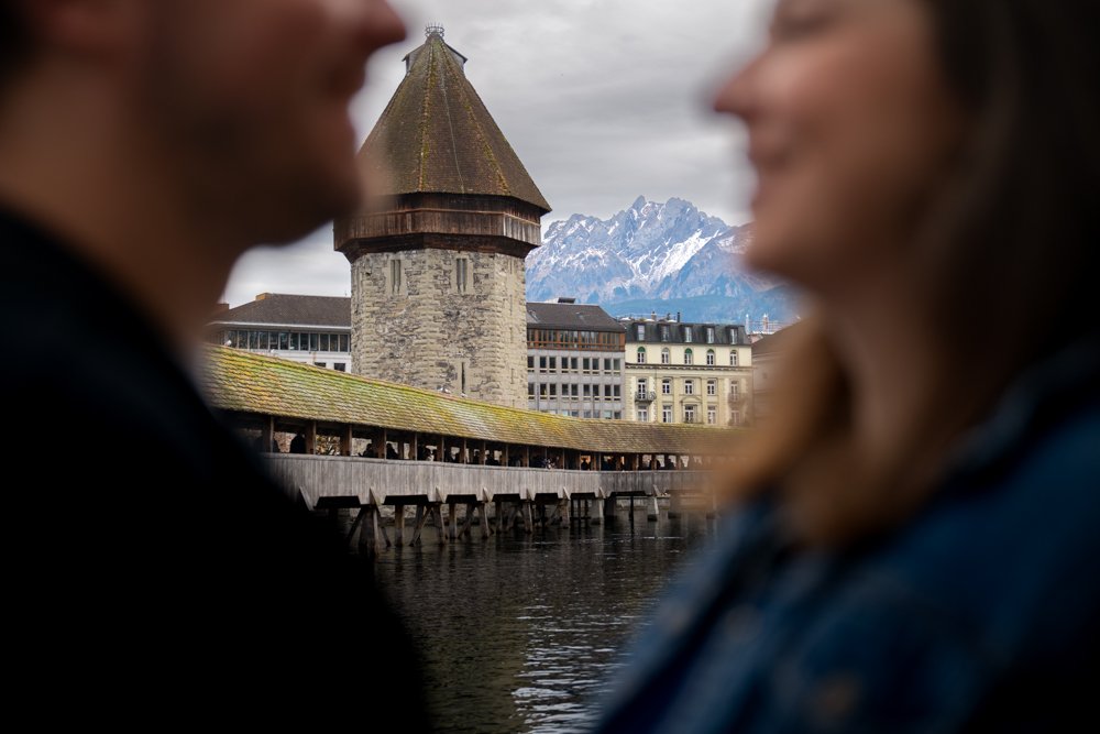 Zwei Menschen stehen vor einer historischen Brücke mit mittelalterlichem Turm, im Hintergrund Berge, vermutlich in einer Stadt am Fluss in der Schweiz.