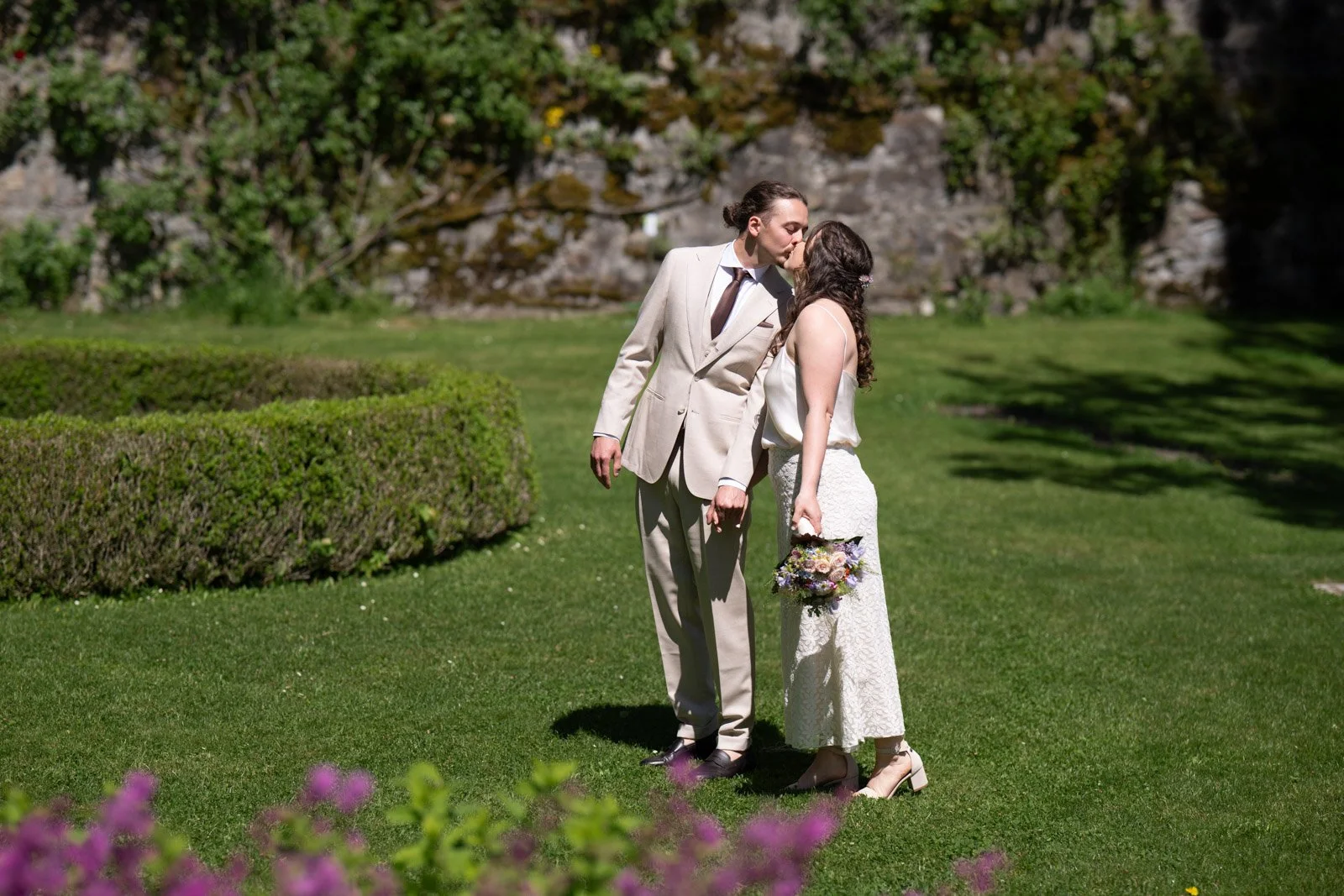 Ein heiratendes Paar küsst sich im Garten bei Sonnenschein, die Frau hält einen Blumenstrauß, beide sind elegant gekleidet, umgeben von grünen Büschen und bunten Blumen.