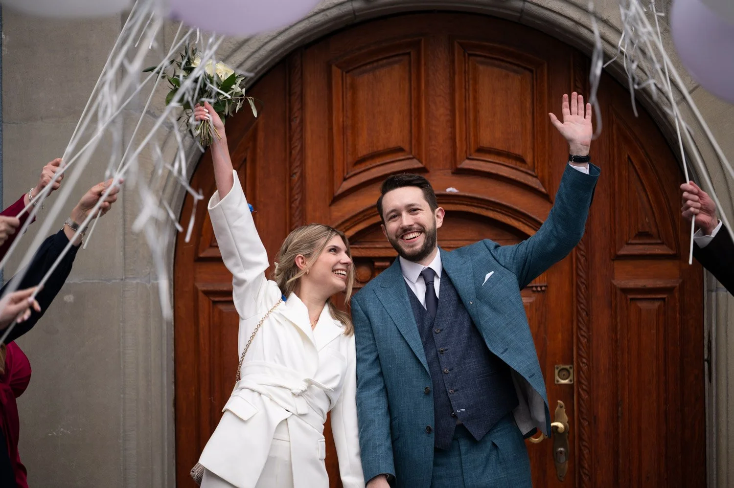 Ein glückliches Brautpaar, das aus einer Kirche mit einem hölzernen Tor tritt, umgeben von Hochzeitsgästen, die Blumen und Luftballons halten.