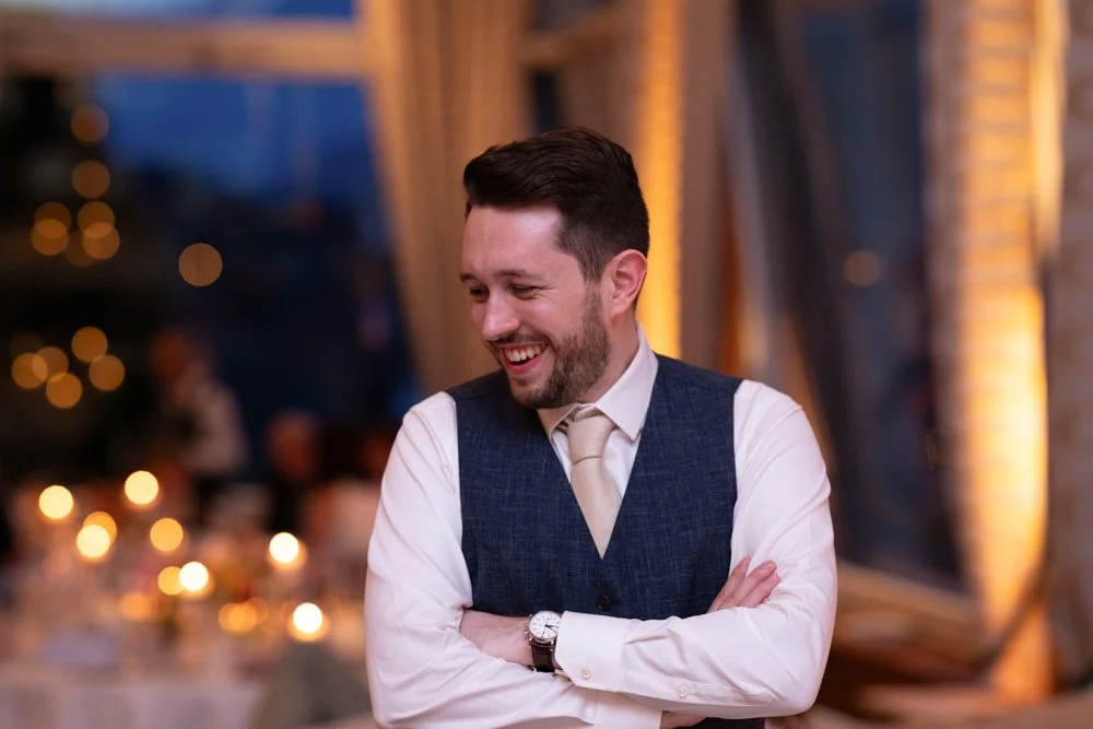 Ein lachender Mann mit dunklem Haar und Bart, trägt eine weiße Hemd und eine dunkle Weste, steht in einem festlich dekorierten Raum bei einer Veranstaltung.