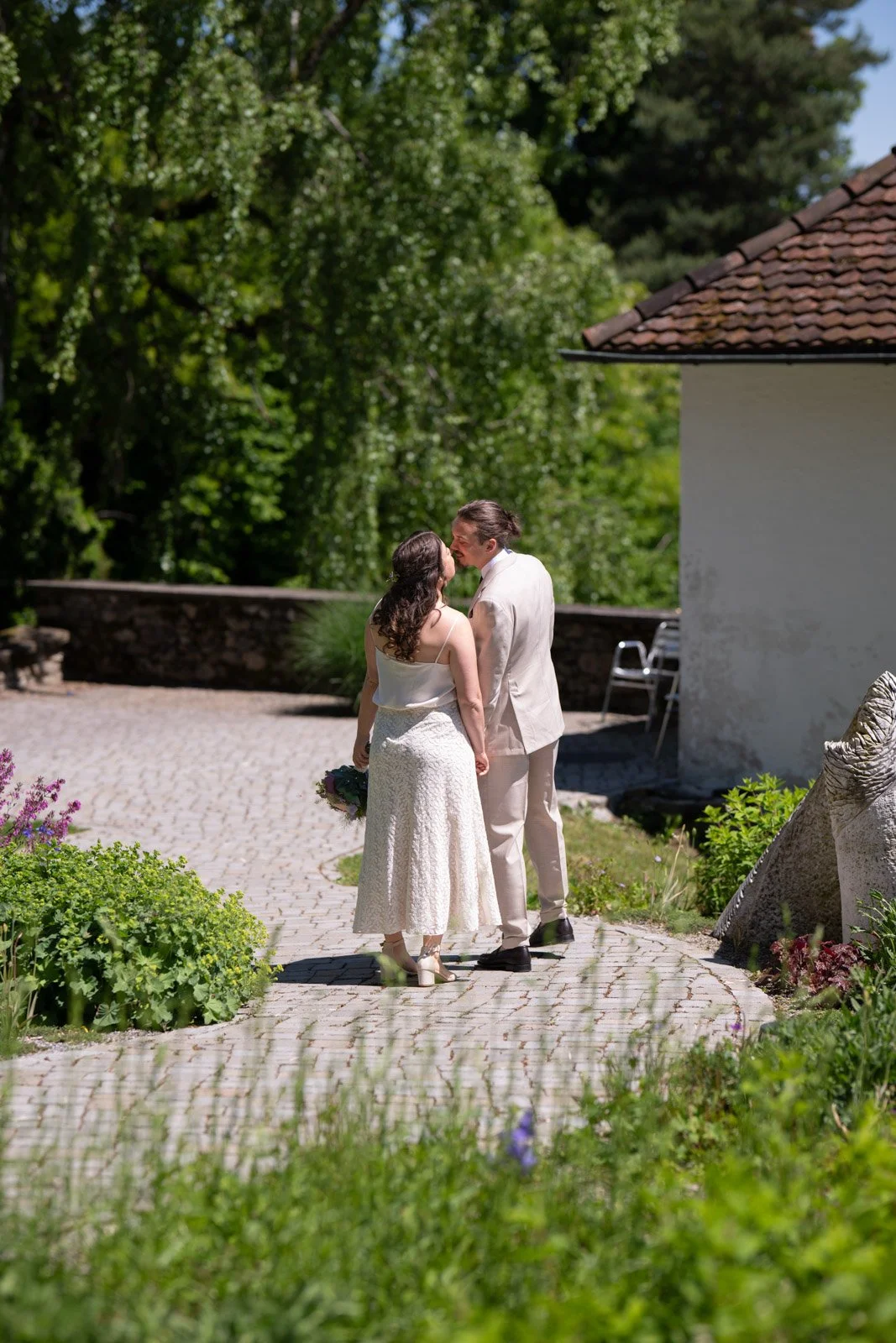 Ein heiratendes Paar steht im Garten und küsst sich an einem sonnigen Tag.
