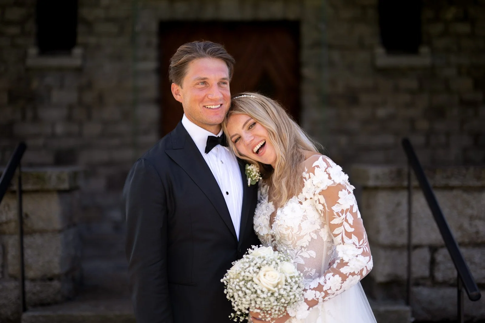 Ein lachendes Brautpaar steht vor einer alten Steinmauer, der Bräutigam trägt einen schwarzen Anzug mit Fliege, die Braut trägt ein langärmeliges weißes Hochzeitskleid mit Blumenmuster und hält einen Brautstrauß aus weißen Blumen.