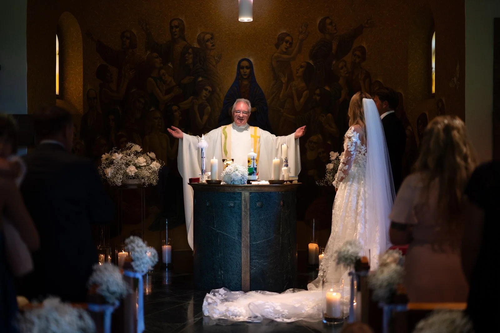 Ein Priester segnet ein Brautpaar während einer Hochzeit in einer Kirche, im Hintergrund eine religiöse Wandmalerei.