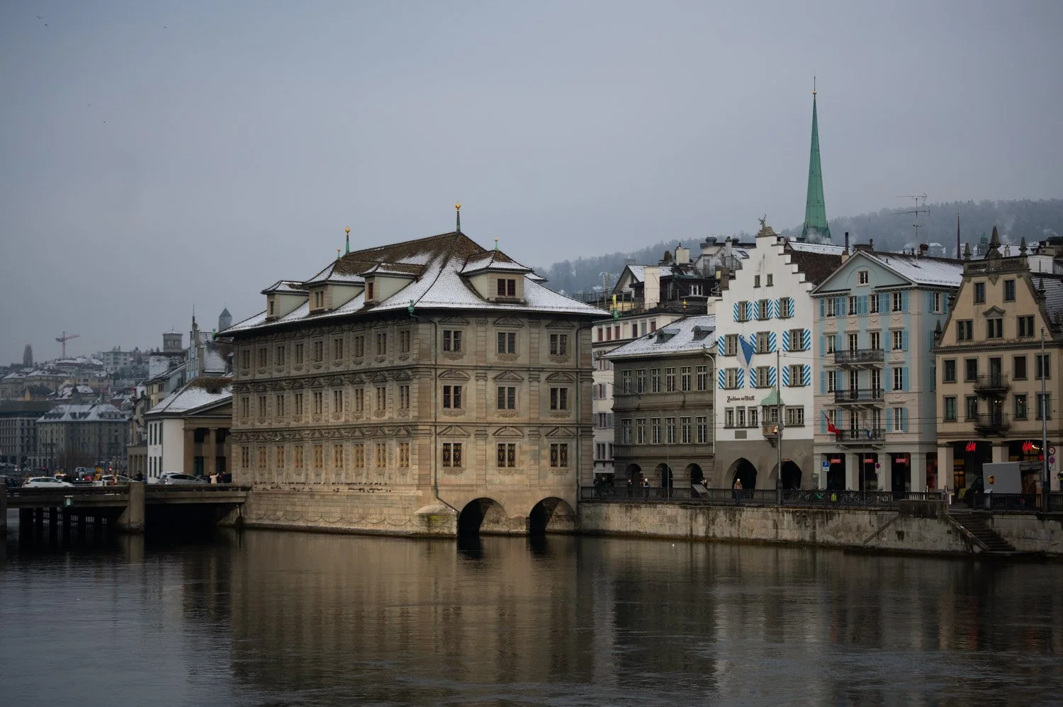 Historische Gebäude am Fluss mit verschneiten Dächern, im Hintergrund Berge und Wolken, Stadtansicht von Zürich