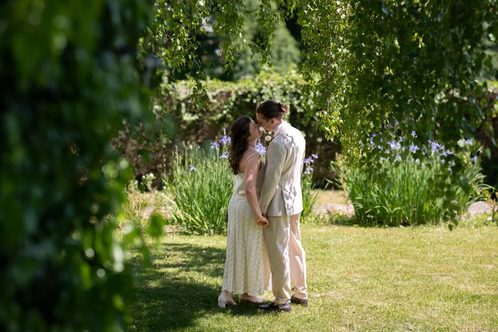 Ein Paar steht sich in einem Garten gegenüber, hält Händchen und schaut sich liebevoll an. Es ist tagsüber, der Garten ist grün und blüht. Die Szene wirkt romantisch und idyllisch.