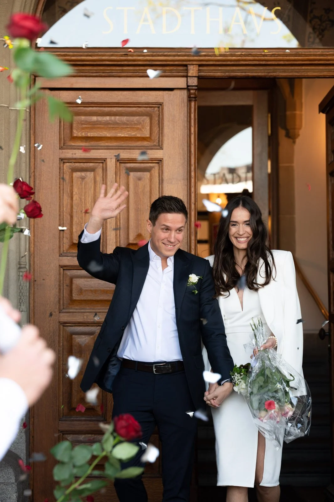 Ein frisch verheiratetes Paar verlässt freudestrahlend ein Gebäude, umgeben von Konfetti und Rosen, während sie sich die Hände halten. Das Paar trägt elegante Kleidung, die Frau hält einen Blumenstrauß. Im Hintergrund ist ein Holzportal mit der Aufsc