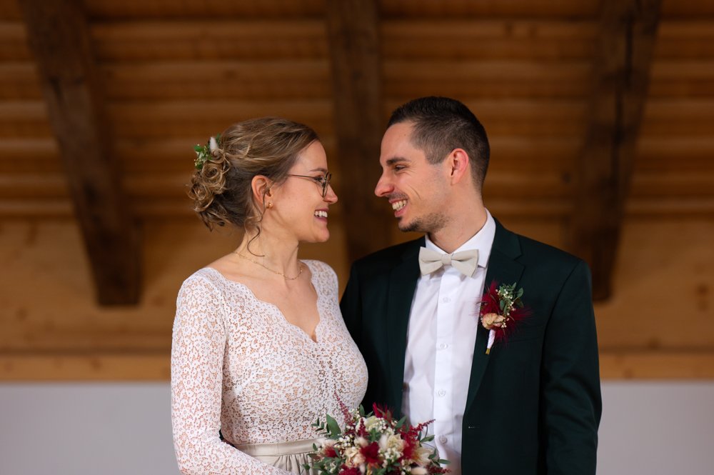 Ein Brautpaar bei einer Hochzeit, die Braut trägt ein weißes Kleid mit Spitze und hält einen Blumenstrauß, der Bräutigam trägt einen schwarzen Anzug mit weißem Hemd und Fliege, beide lächeln sich an.