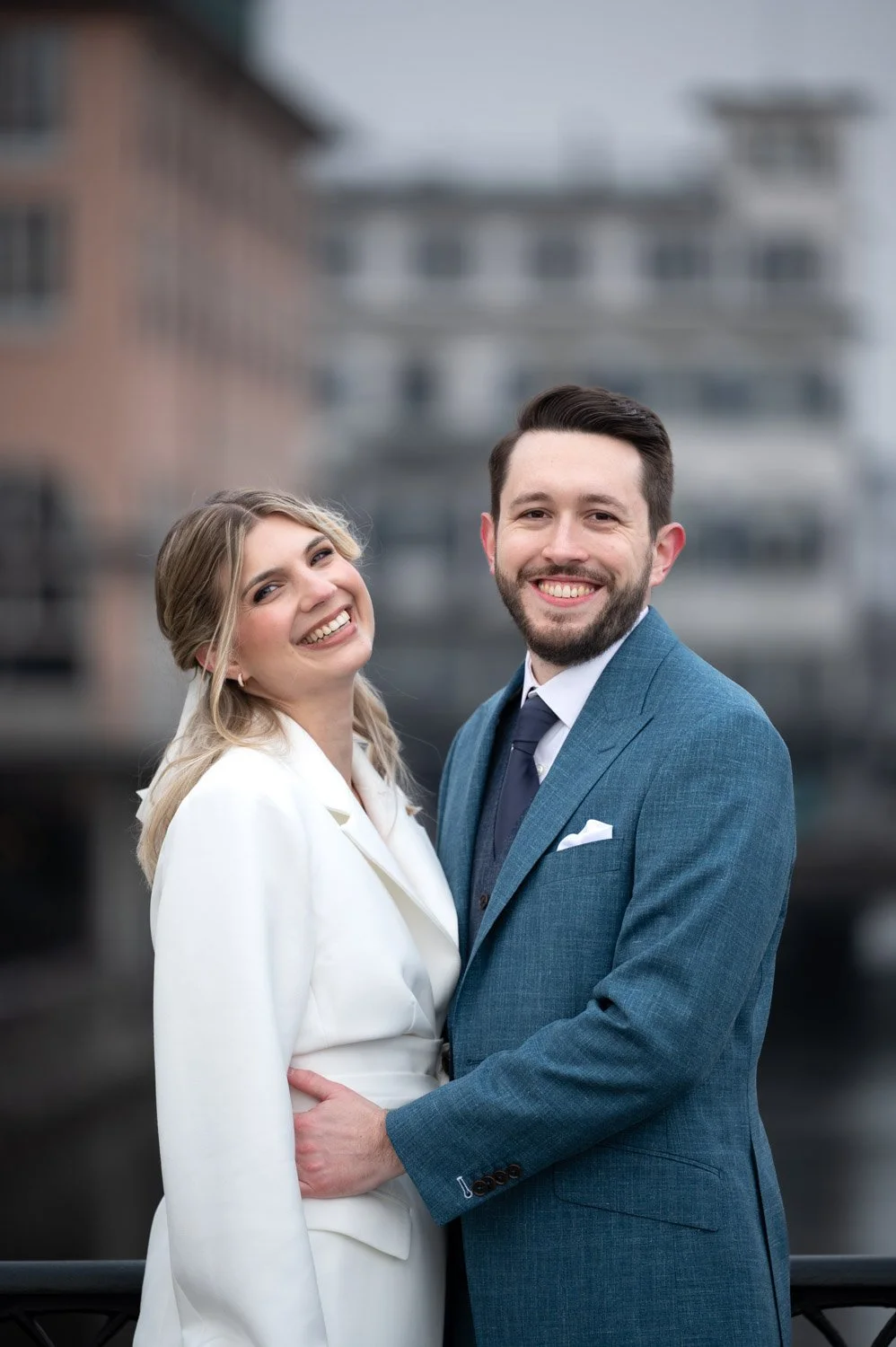 Ein lachendes Paar in formeller Kleidung, die im Freien vor einem städtischen Hintergrund posieren.