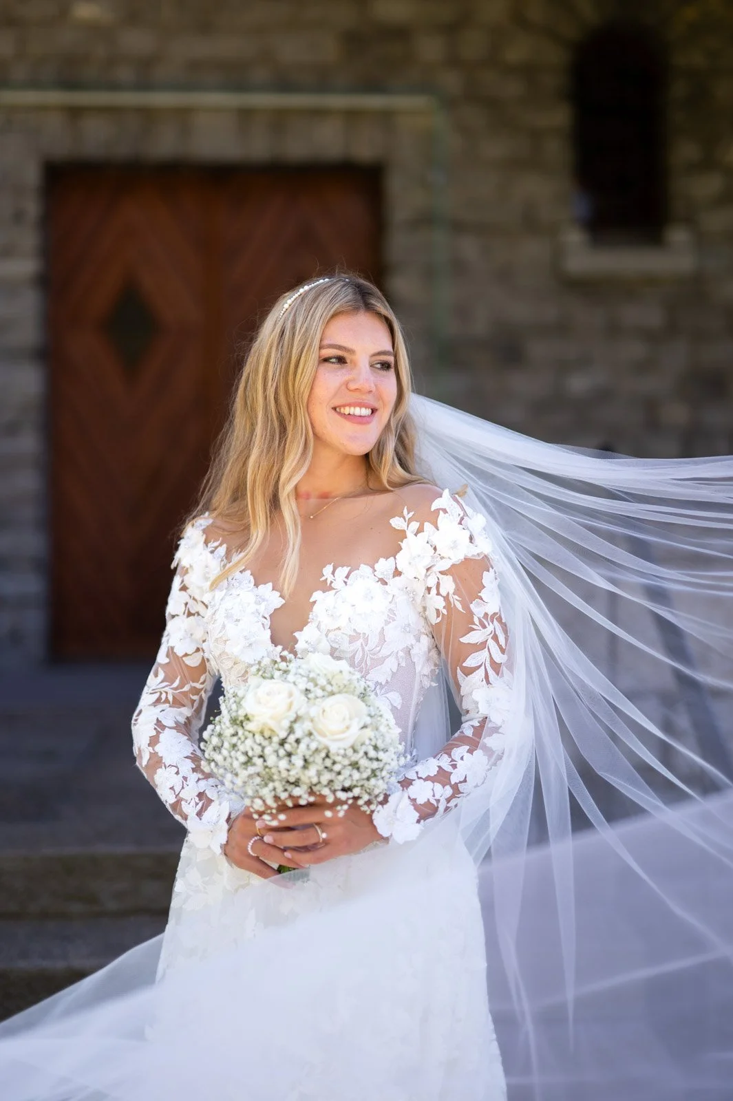 Braut in weißem Hochzeitskleid mit Blumenmuster und Schleier hält einen Blumenstrauß aus weißen Rosen und Babybreäucher, steht vor einer Holz- und Steinwand.