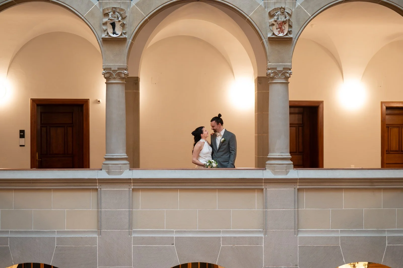 Ein brautpaar steht sich in einem historischen Gebäude gegenüber, umgeben von klassischen Steinbögen und Säulen, mit warmem Licht im Hintergrund.
