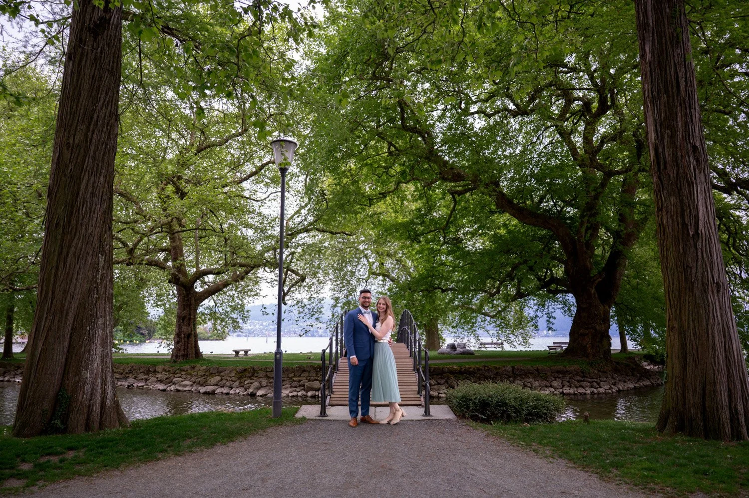 Ein Paar in formeller Kleidung steht auf einer kleinen Brücke in einem parkähnlichen Bereich mit großen Bäumen, einem Wasserlauf und Sitzbänken im Hintergrund.