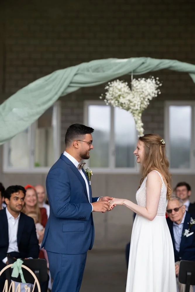 Braut und Bräutigam während der Hochzeitszeremonie, die sich die Hände halten und in die Augen schauen, umgeben von Freunden und Familie in einem festlich geschmückten Raum.
