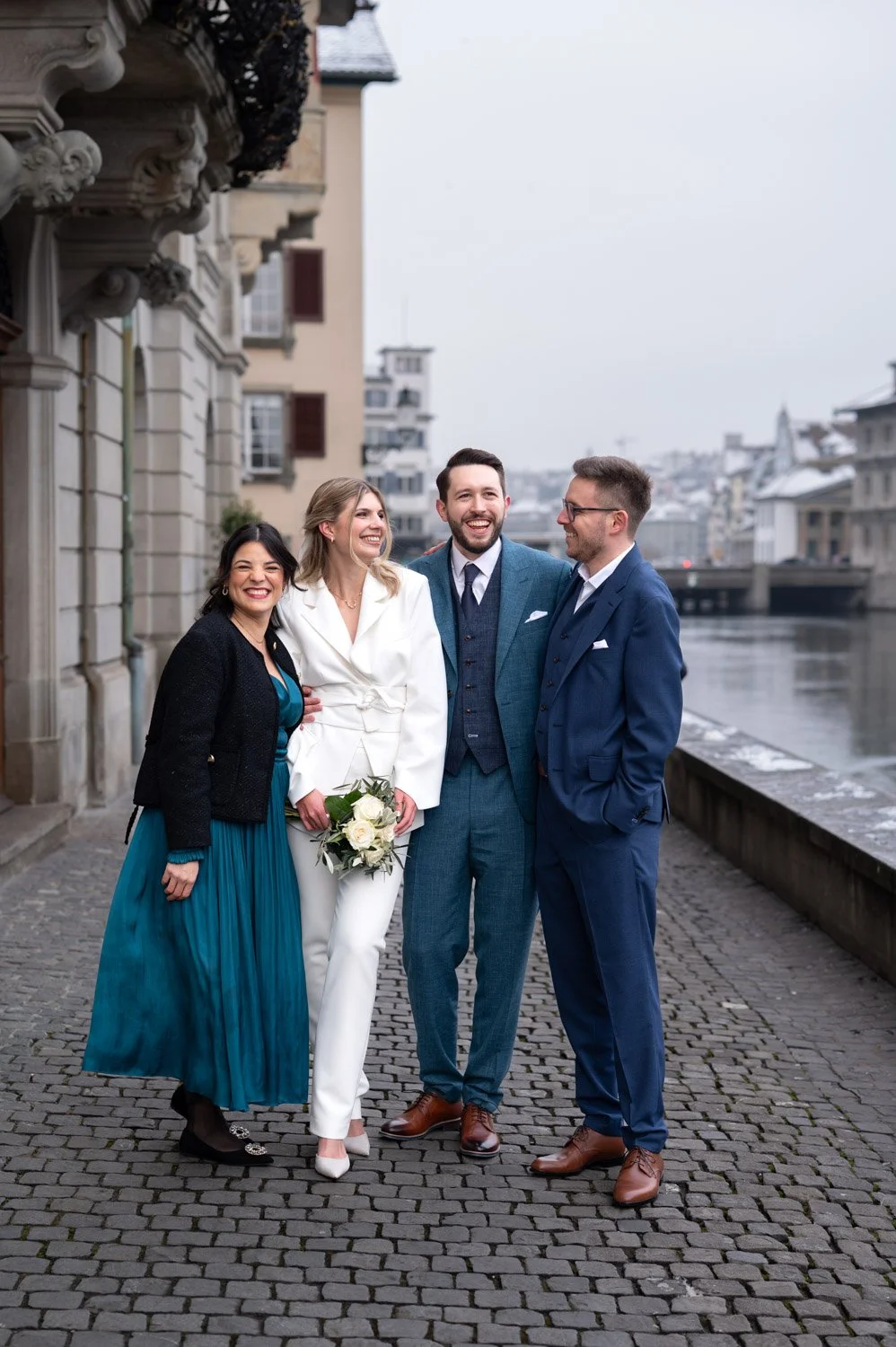 Vier Menschen in formeller Kleidung stehen lächelnd am Wasser in einer Stadt mit alten Gebäuden und Brücke im Hintergrund.