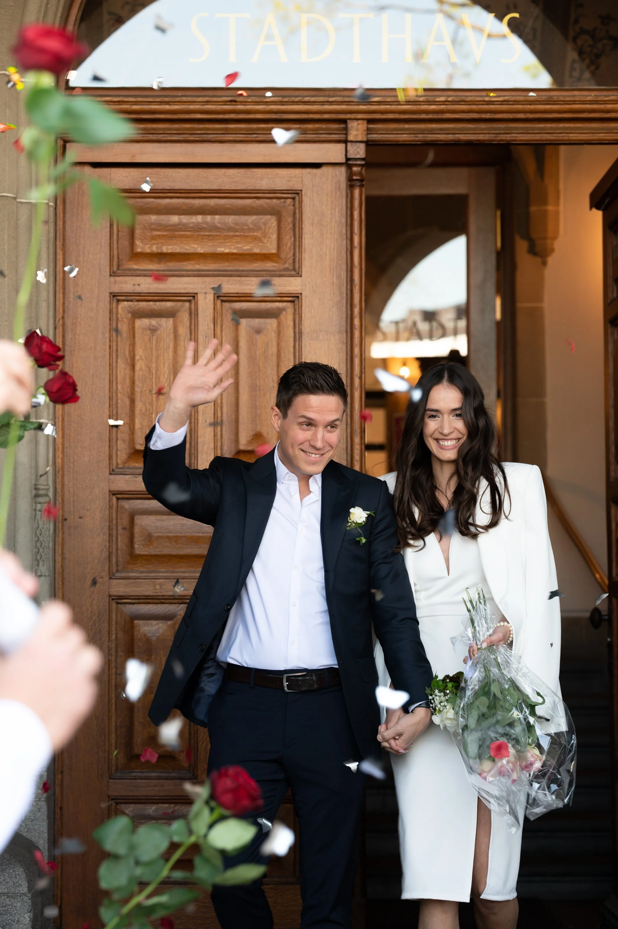 Ein frisch verheiratetes Paar verlässt das Stadthaus in Zürich, umgeben von spaltenden Rosen und konfettibstreuenden Gästen, während sie Händchen halten und lächeln.